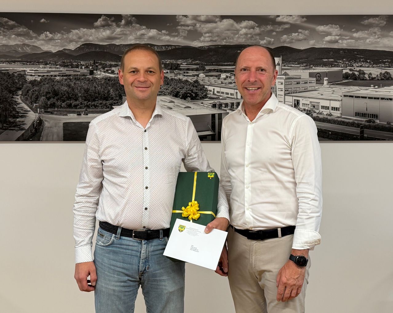Zwei Männer stehen nebeneinander, einer hält eine Geschenkbox mit gelber Schleife und lächelt, vor einem Foto mit einem industriellen Hintergrund.