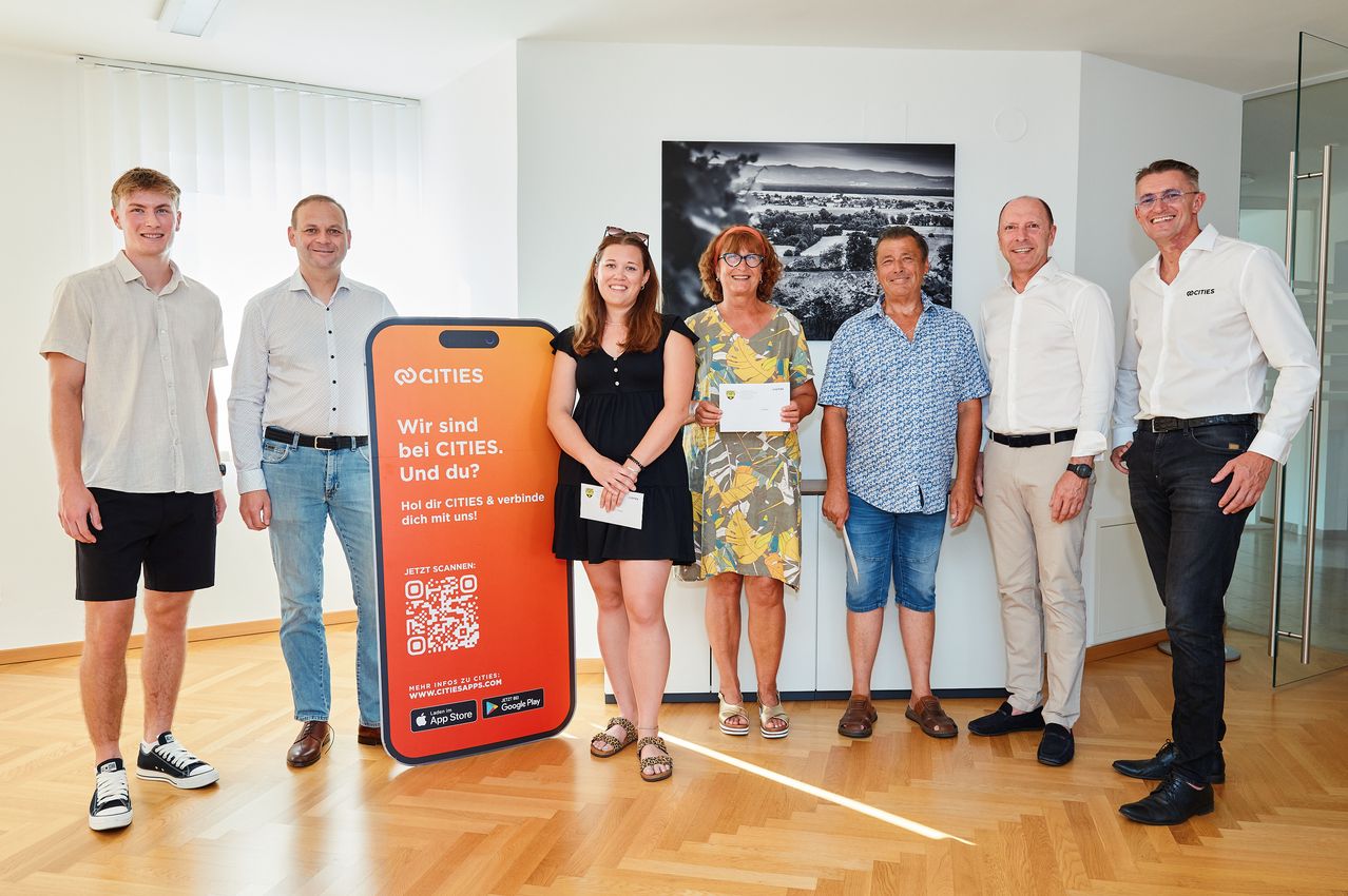 Fünf Menschen stehen lächelnd in einem Raum. Eine Frau hält ein Zertifikat. Ein orangefarbenes Poster mit Text und QR-Code steht vor ihnen.
