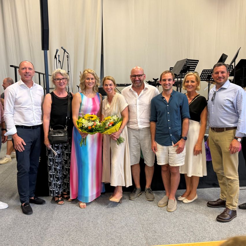 Eine Gruppe von Menschen steht zusammen und lächelt für ein Foto. Eine Frau in der Mitte hält Blumen. Dahinter sind Musikinstrumente und eine Bühne zu sehen.