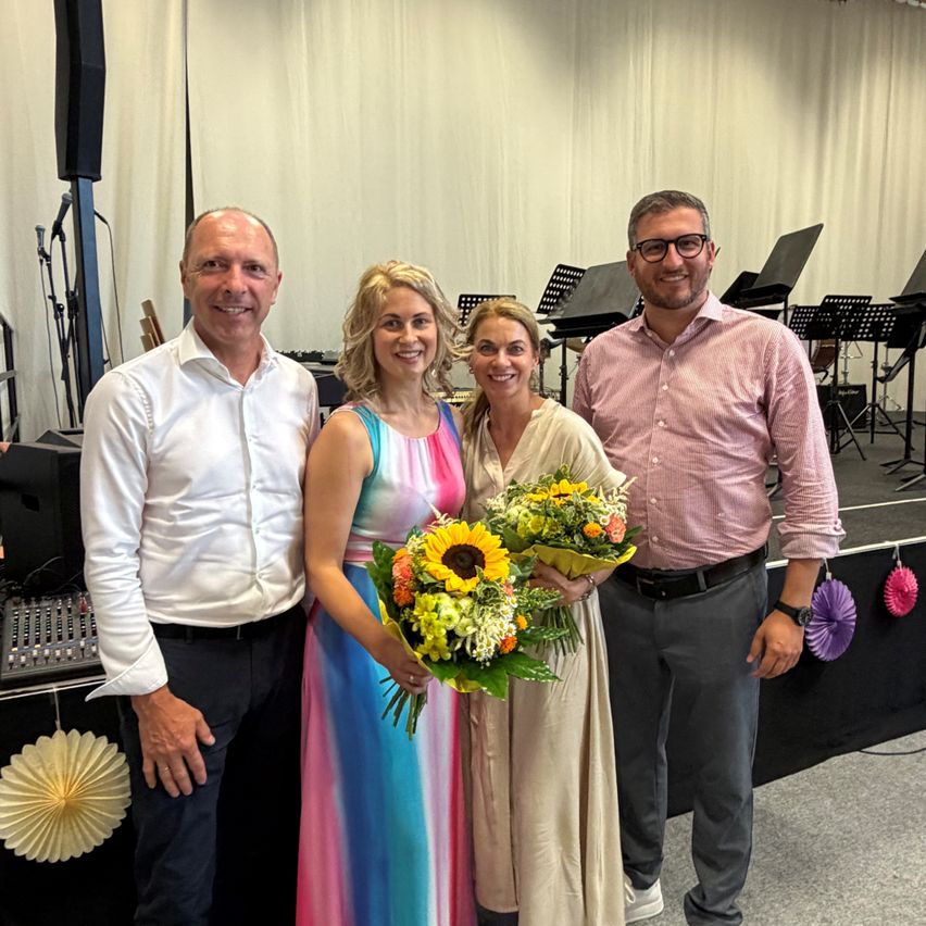 Drei Personen stehen in einem Raum mit Musikinstrumenten und einer Bühne im Hintergrund. Eine Frau hält Blumen und lächelt mit zwei Männern.