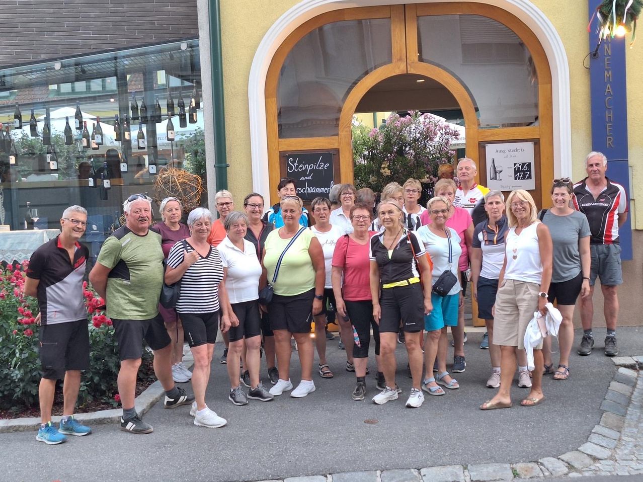 Eine Gruppe von Erwachsenen in verschiedenen Farben von Shirts und Shorts posiert vor einem Geschäft mit einem Schild auf Deutsch, einer Tafel und einer Blumendekoration.