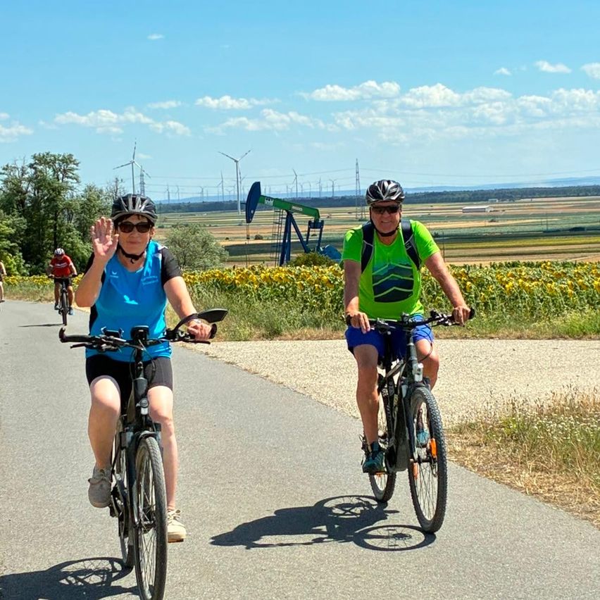 Eine Gruppe von Radfahrern fährt auf einer Straße mit Sonnenblumen und Windrädern im Hintergrund. Ein Radfahrer winkt.
