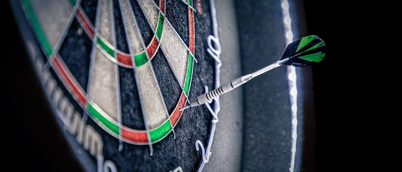 Eine Nahaufnahme eines Dartboards mit einem eingesteckten Dart. Das Dartboard hat einen schwarzen Rand mit weißen Zahlen und farbigen Bereichen. Der Dart ist silbern mit einer grünen und schwarzen Spitze.