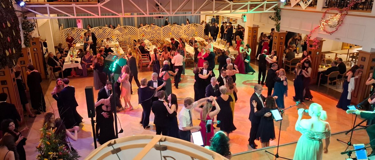 Eine obere Ansicht eines Ballsaales mit tanzenden Menschen, einige stehend, und einer Bühne mit einer Sängerin und einer Band, umgeben von Tischen mit Blumen und Lichtern.
