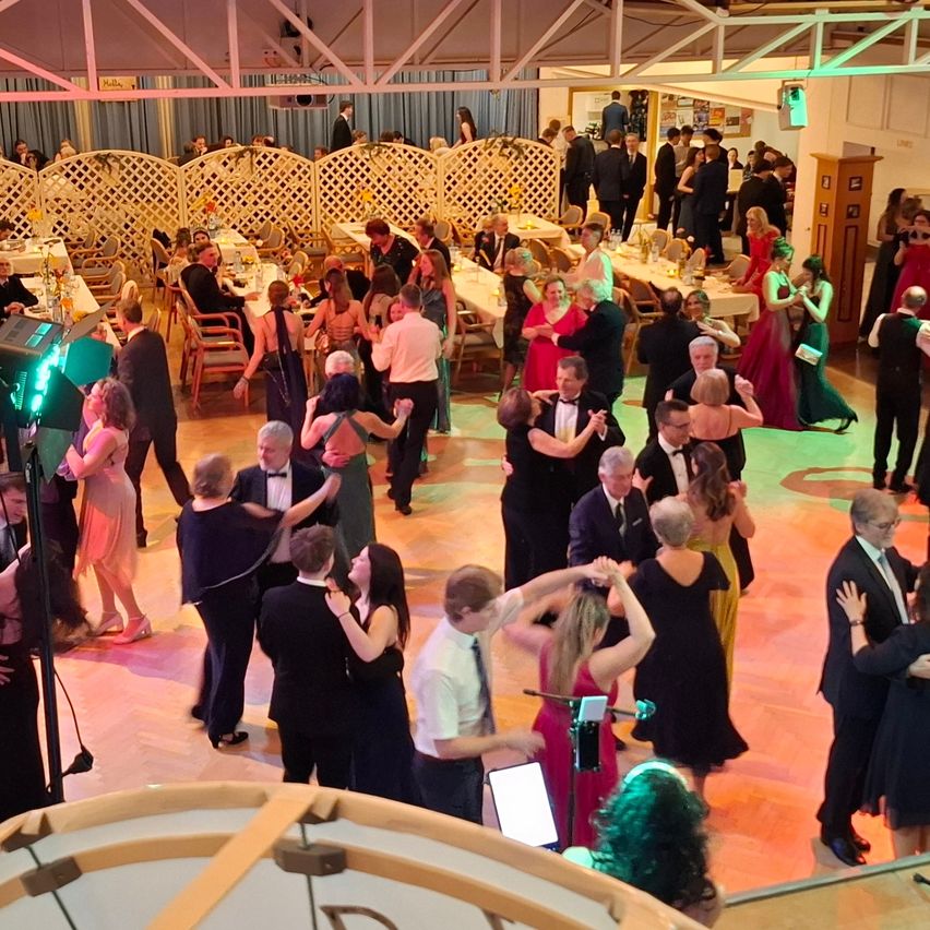 Eine obere Ansicht eines Ballsaales mit tanzenden Menschen, einige stehend, und einer Bühne mit einer Sängerin und einer Band, umgeben von Tischen mit Blumen und Lichtern.