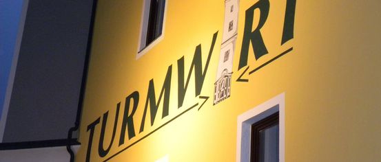 A yellow building with the words Turmwirt and an arrow pointing to a tower logo on the right. Three windows with white frames and black outlines.