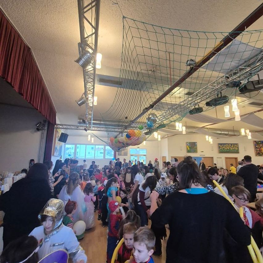 Viele Kinder und Erwachsene in Kostümen versammeln sich in einer Halle, wahrscheinlich für eine Halloween-Party. Einige tragen Brillen, Hüte und Masken. Es hängen Netze und Lichter.