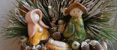 Eine Keramik-Krippe in einem Topf mit Maria, Josef und dem Baby Jesus, umgeben von Muscheln, Tannenzapfen und Grün. Der Topf ist mit vereisten Zweigen dekoriert.