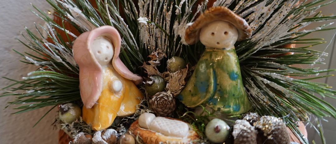Eine Keramik-Krippe in einem Topf mit Maria, Josef und dem Baby Jesus, umgeben von Muscheln, Tannenzapfen und Grün. Der Topf ist mit vereisten Zweigen dekoriert.