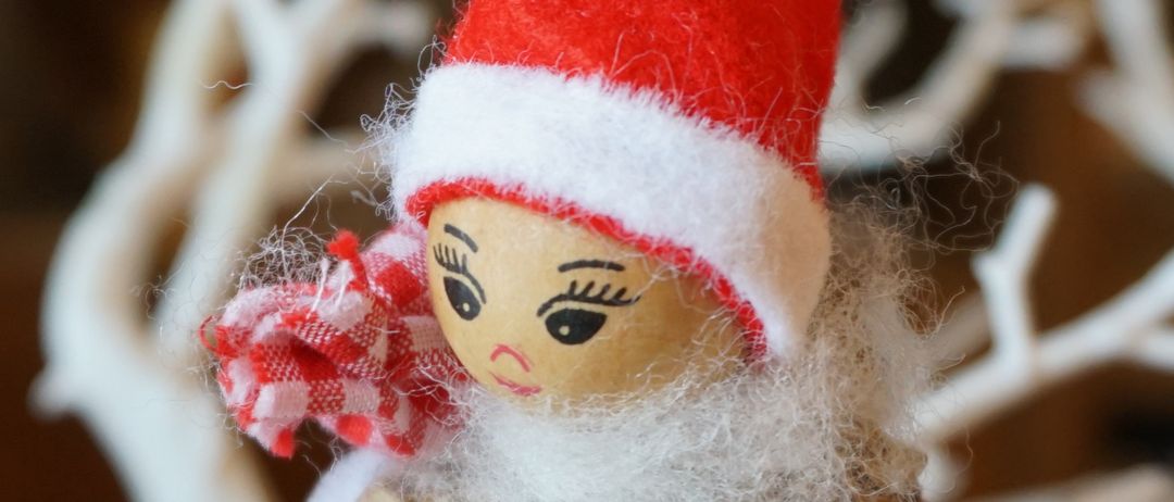 Eine kleine, handgefertigte Elbenpuppe mit rotem Hut und weißem Bart, die einen rot-weiß karierten Schal trägt und auf einer Basis aus Tannenzapfen sitzt.