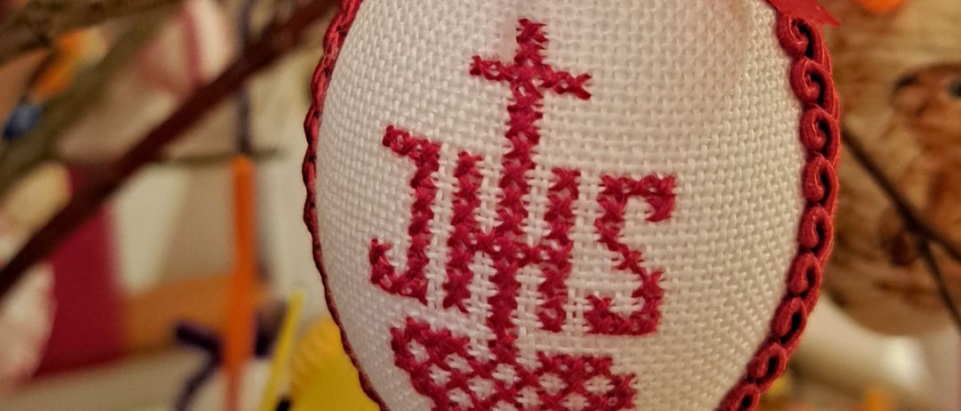 Ein rot-weißes Ornament mit den Buchstaben JHS in Rot gestickt und einem Kreuz oben, gebunden mit einem roten Band.