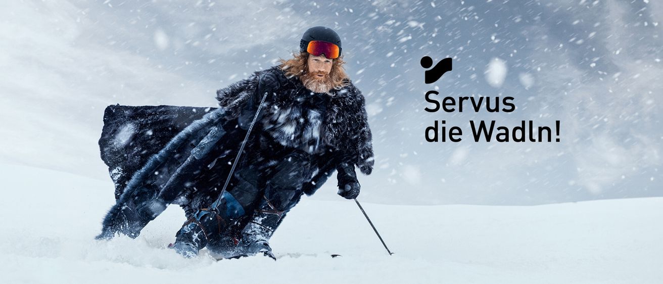 Ein Skifahrer mit Schutzbrille und Pelzmantel skisrt im Schnee. Ein Logo und der Schriftzug 'Server die W' befinden sich in der oberen rechten Ecke.
