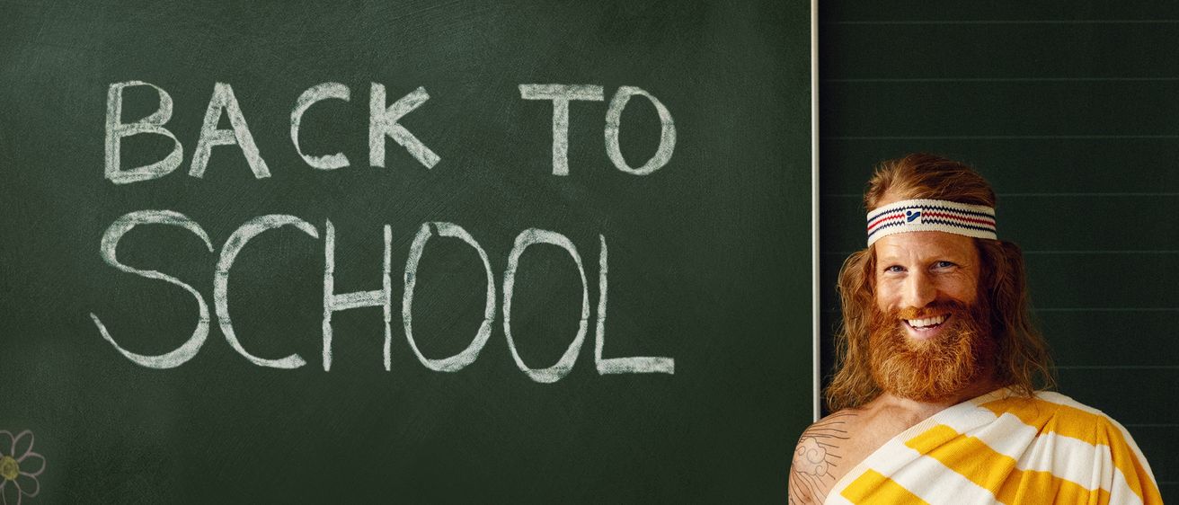 Ein Mann mit Bart und Tätowierungen steht vor einer Tafel mit den Worten 'ZURÜCK ZUR SCHULE'. Er trägt einen gelben und weißen Umhang und eine Kopfband.