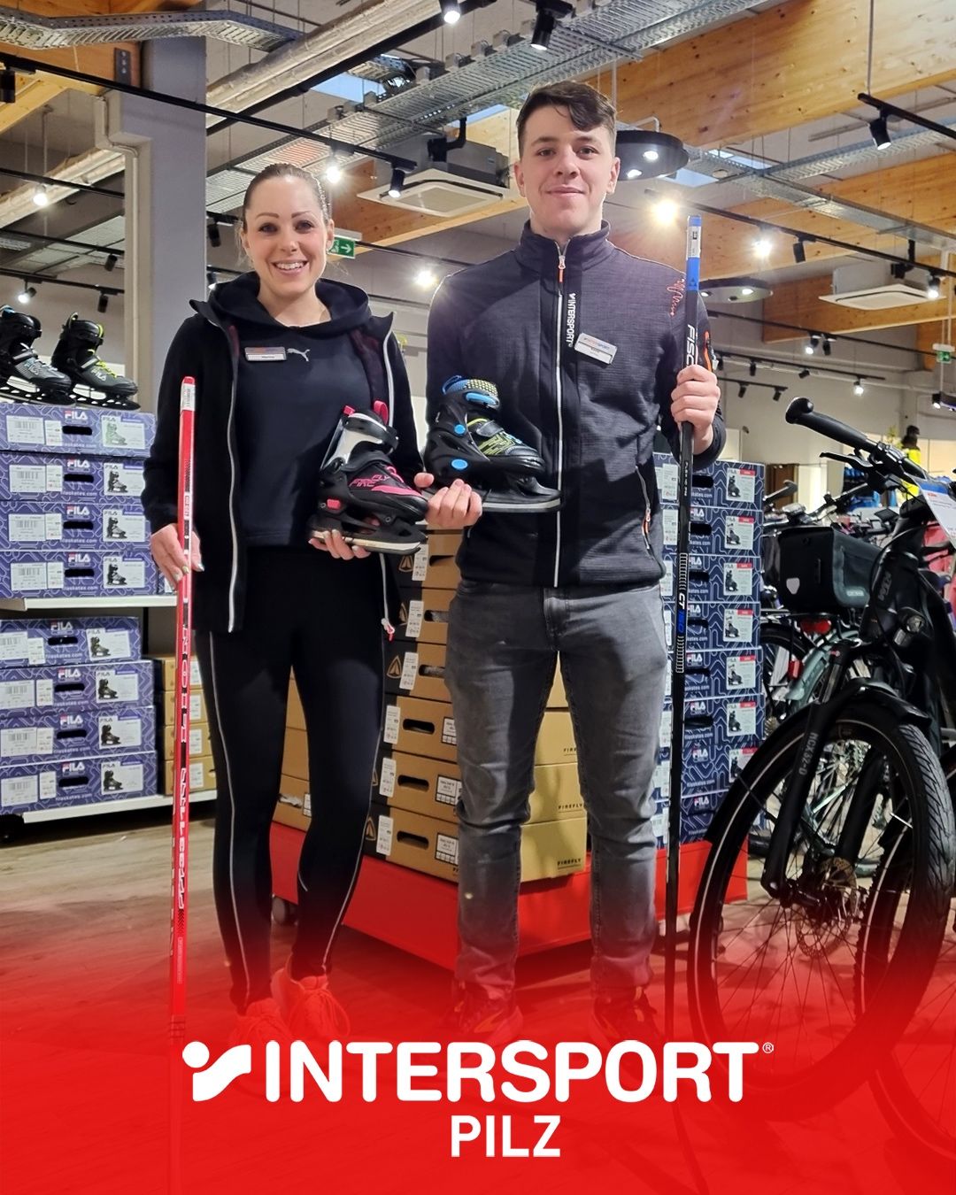 Zwei Personen stehen in einem Geschäft mit Sportausrüstung. Die Frau hält ein Paar Rollschuhe und einen Skistock. Der Mann hält einen Skistock und Schlittschuhe. Hinter ihnen sind Fahrräder und Sportgeräte zu sehen.