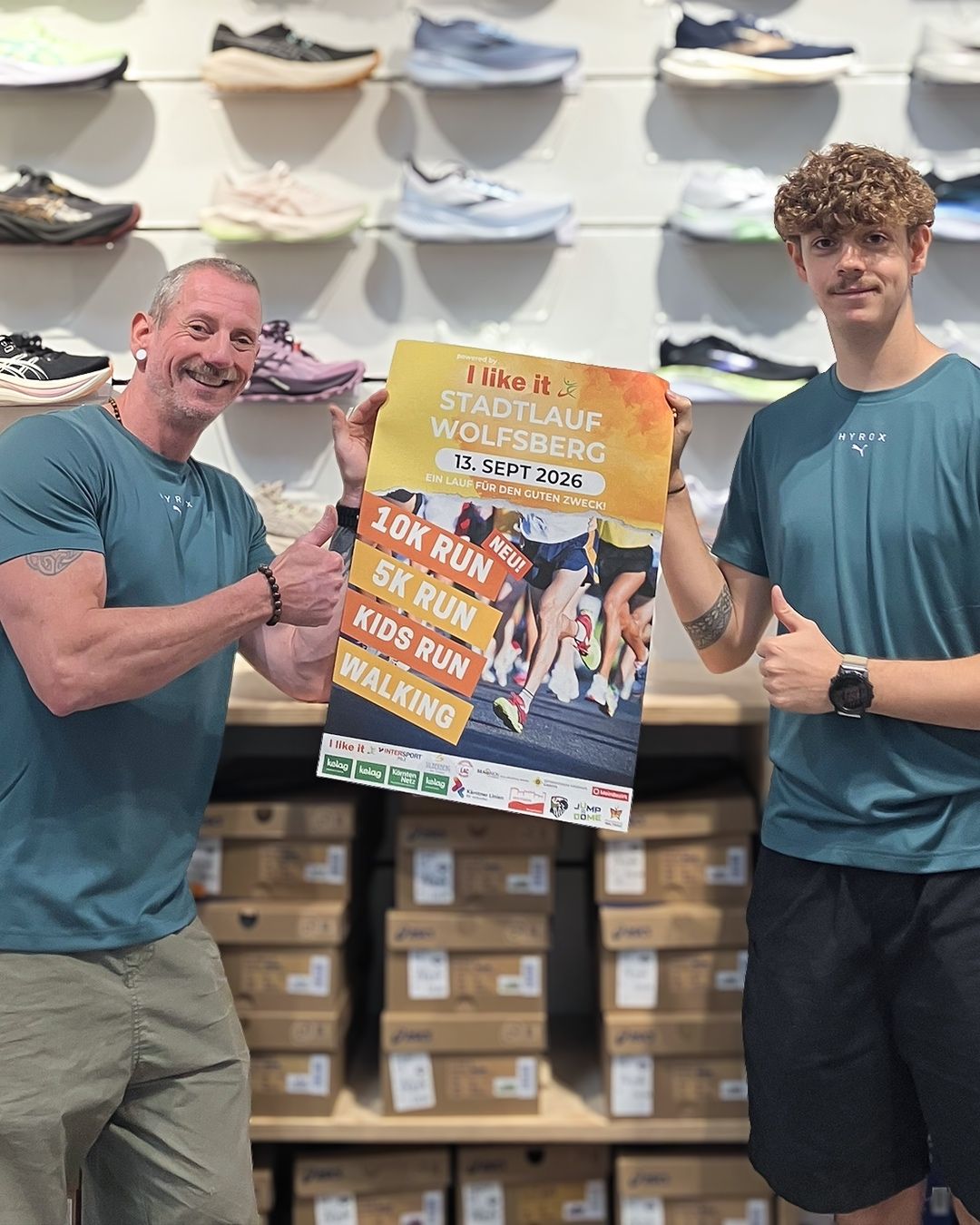 Zwei Männer in einem Schuhgeschäft, einer hält ein Plakat für einen Laufwettbewerb in Wolfsberg am 13. September 2026.