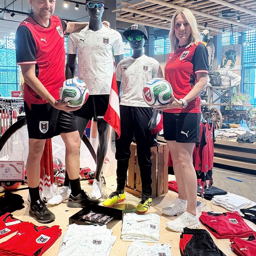 Three people stand in a clothing shop with sports jerseys and soccer balls. The two women hold balls and the man smiles. Mannequins and sports gear are in the background.