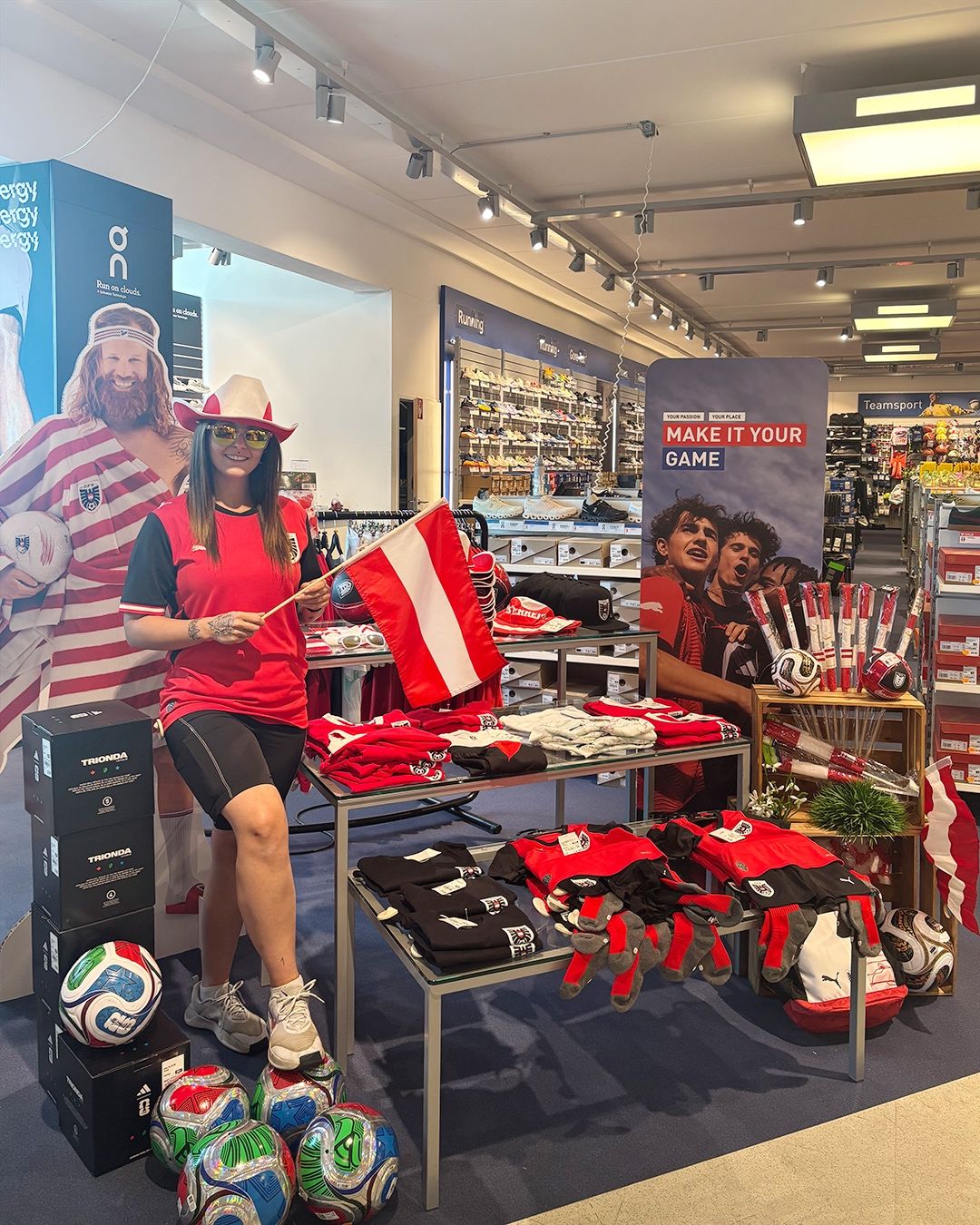 Eine Frau hält eine Flagge in einem Sportgeschäft, mit Merchandise und einem großen Plakat mit der Aufschrift 'MAKE IT YOUR GAME'. Sportausrüstung und Trikots sind auf Tischen ausgestellt.