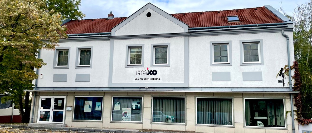 Ein weißes Gebäude mit roten Dachziegeln, mehreren Fenstern und einem Schild, das 'HCK Gas Wasser Heizung' besagt. Die Vorderseite hat Glasfenster und Türen. Ein Baum steht auf der linken Seite.