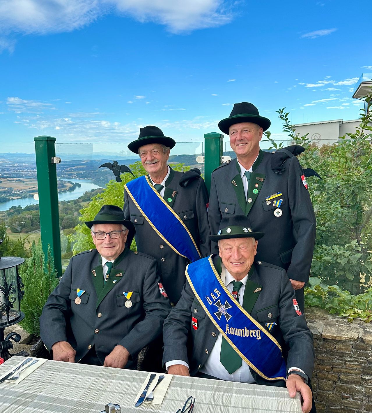 Vier Männer in traditionellen Uniformen mit Medaillen und Schärpen, die vor einem Tisch sitzen, mit einem malerischen Blick auf einen Fluss und Berge im Hintergrund.