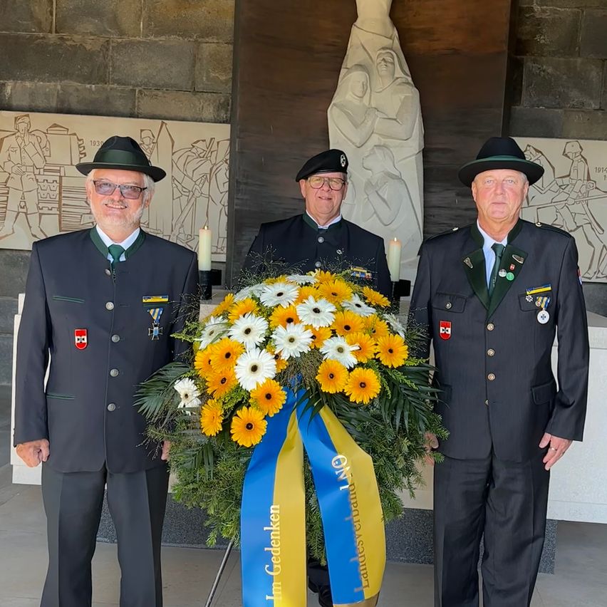Drei Männer in Militäruniformen stehen neben einem Blumenarrangement mit einem blau-gelben Band. Dahinter befindet sich eine große Steinmauer mit einer Statue.