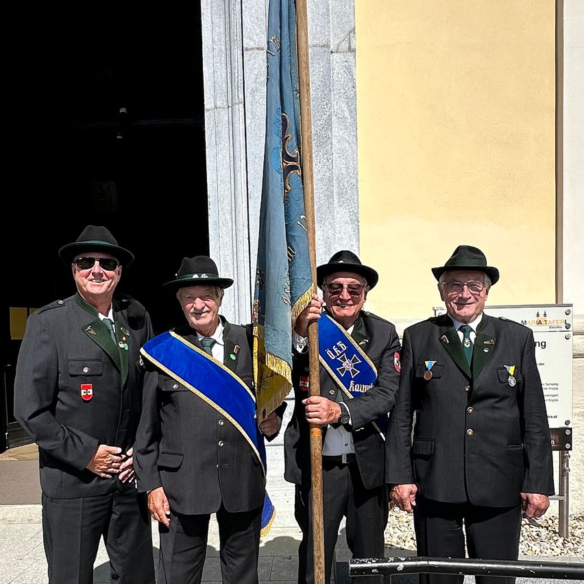 Vier Männer in Uniformen stehen lächelnd draußen, einer hält eine Flagge. Sie stehen vor einem Gebäude mit gelber Wand.