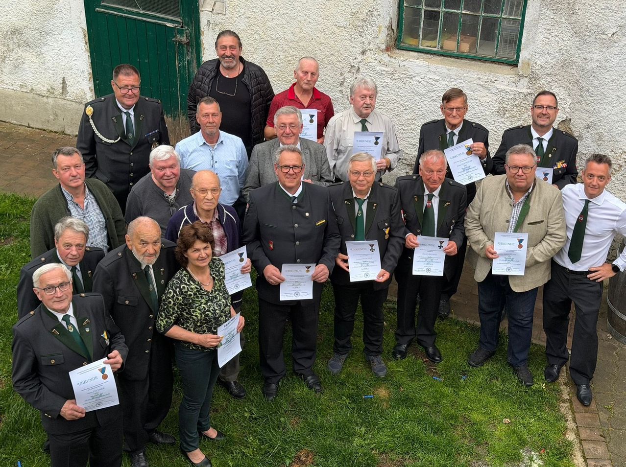 Eine Gruppe von Männern und Frauen in formeller Kleidung, einschließlich Uniformen und Krawatten, versammelt sich vor einem Gebäude für ein Foto. Sie halten Zertifikate in den Händen.