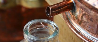 Ein Kupferdestillationsapparat ist zu sehen, mit einem Kupferrohr, das in eine Glasflasche mit Wassertropfen führt. Der Hintergrund ist unscharf mit Kupfergefäßen.