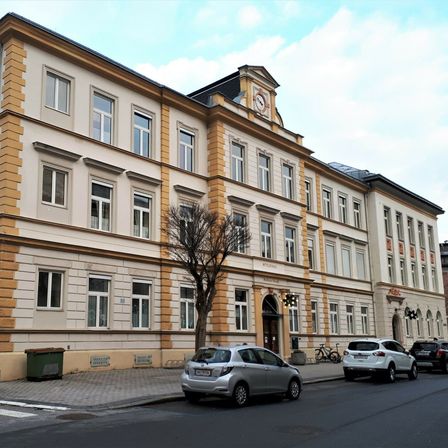 Ein mehrstöckiges Gebäude mit gelben und weißen Akzenten steht neben einer Straße, mit geparkten Autos davor.