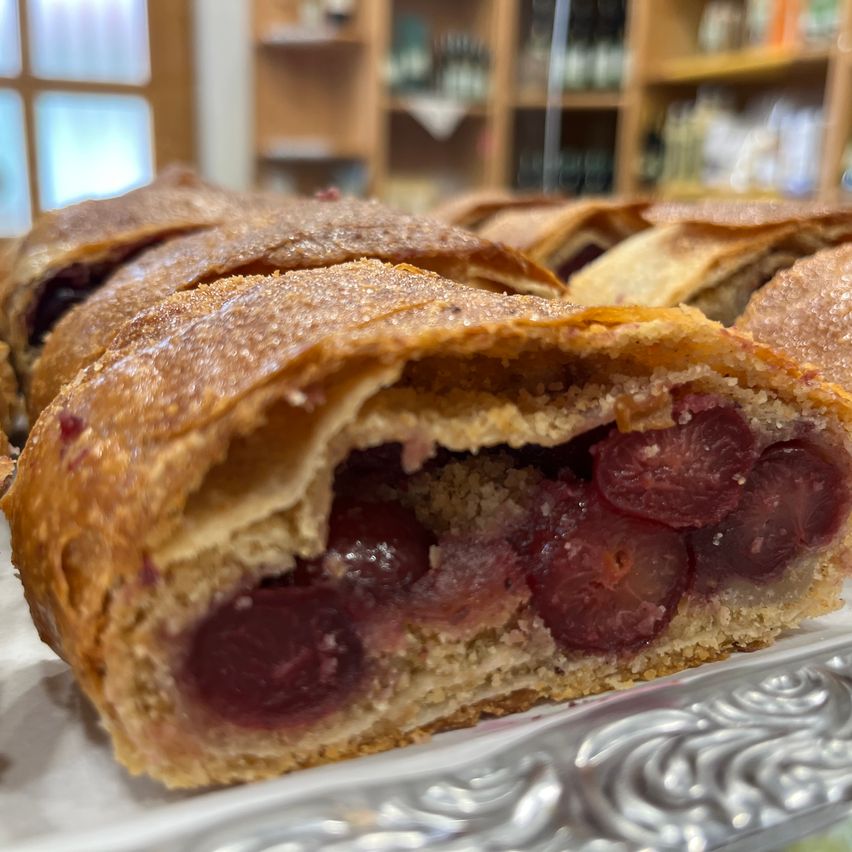 Ein Nahaufnahme von einer Kirschstrudel-Pastete mit einem sichtbaren Stück, das seine Kirschfüllung zeigt. Die Pastete hat eine goldbraune Kruste. Im Hintergrund ist ein Holzregal zu sehen.