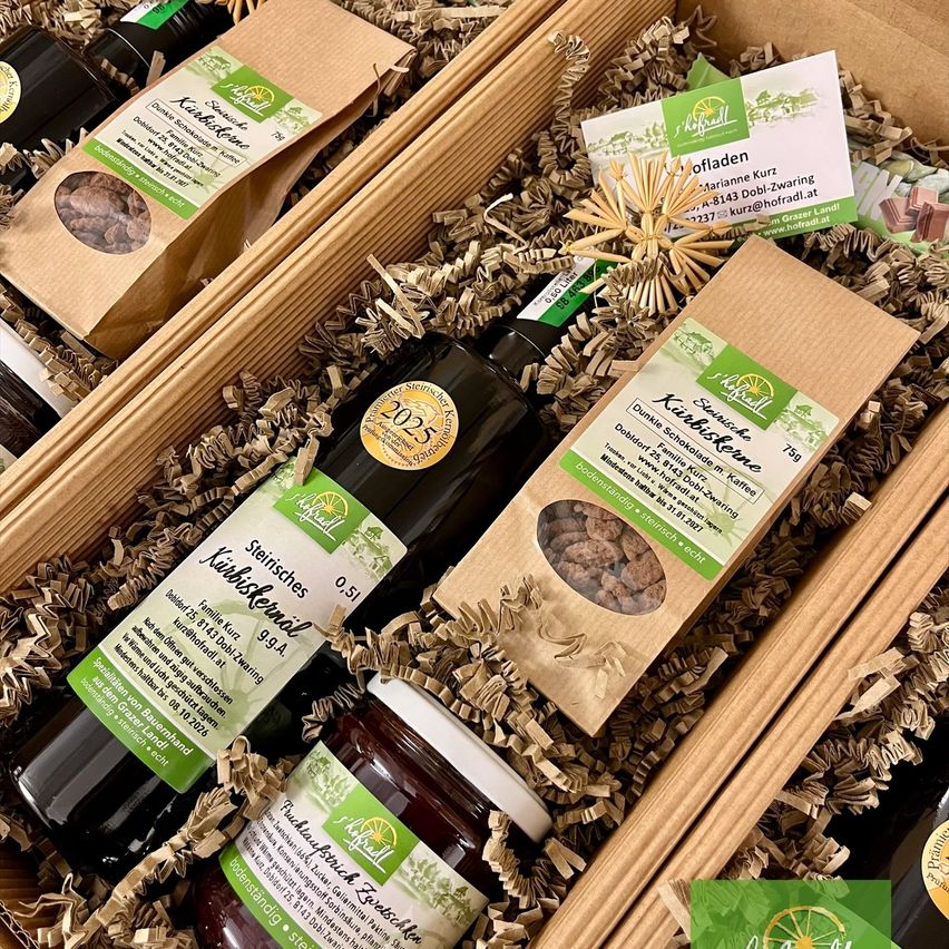 Eine Holzkiste gefüllt mit verschiedenen Produkten, darunter eine Flasche steirisches Kürbiskernöl, ein Glas Fruchtmarmelade und andere Artikel. Die Kiste ist mit zerrissenem Papier gepolstert.