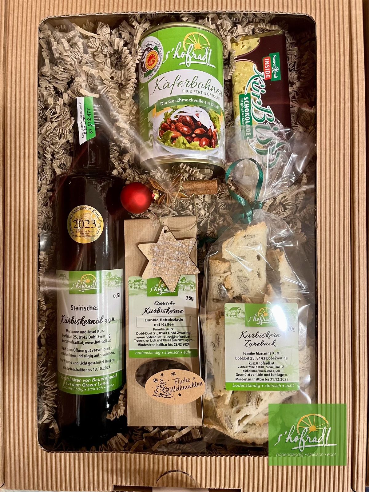 Eine Holzkiste enthält eine Flasche Likör, ein Glas Kaffeebohnen, eine Packung Pralinen und einen Beutel Kekse. Die Flasche ist mit 'Steirische' und 'Kürbiskernöl' beschriftet und hat einen Aufkleber, der angibt, dass sie 2023 hergestellt wurde. Das Glas ist mit 'Kafferbohne' und einem grünen und weißen Etikett versehen. Die Pralinen und Kekse sind in Plastik verpackt und ebenfalls beschriftet.