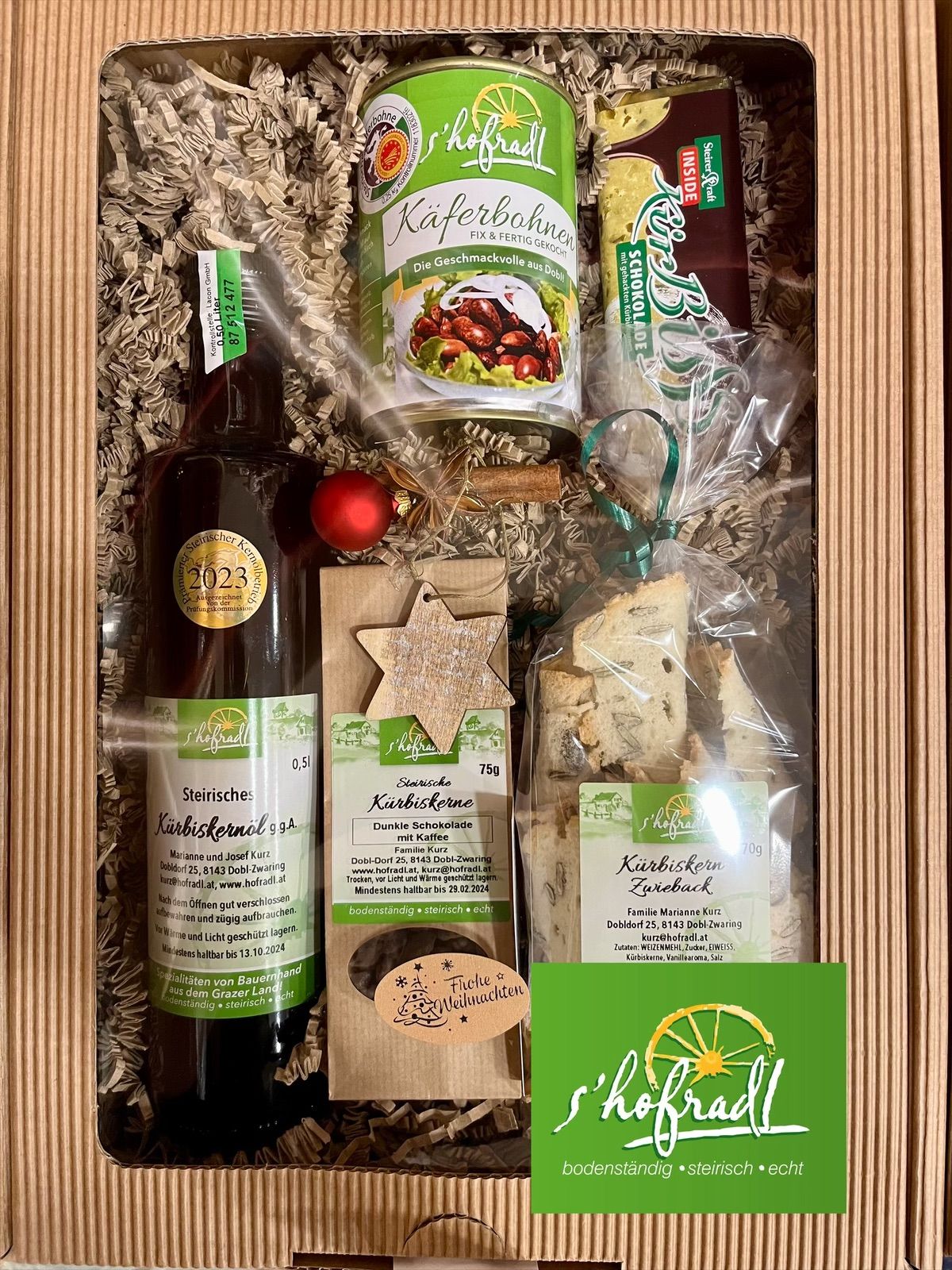 Eine Holzkiste enthält eine Flasche Kaffeelikör, ein Glas Kaffeebohnen, einen Beutel mit schokoladenüberzogenen Keksen, einen Weihnachtsstern, eine Zimtstange und eine rote Weihnachtskugel.