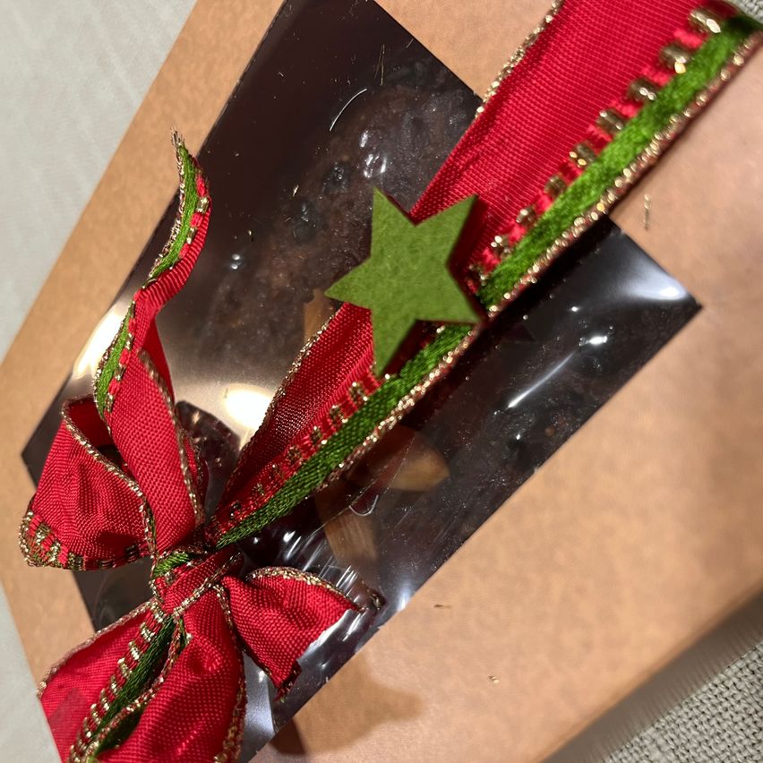 Ein brauner Geschenkkarton aus Papier mit roter Schleife und grünem Stern als Dekoration. Die Schleife ist mit goldenem Glitzer verziert. Der Geschenkkarton enthält eine dunkle Schokoladen-Süßigkeit.