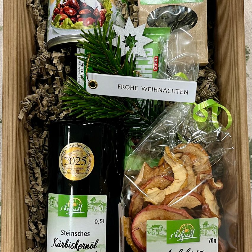 Eine Holzkiste enthält ein Geschenkset für Weihnachten 2025. Es umfasst eine Flasche steirisches Kürbiskernöl, einen Beutel mit getrockneten Apfelscheiben und ein Glas Kirschkompott. Jeder Artikel ist in Cellophan eingewickelt.