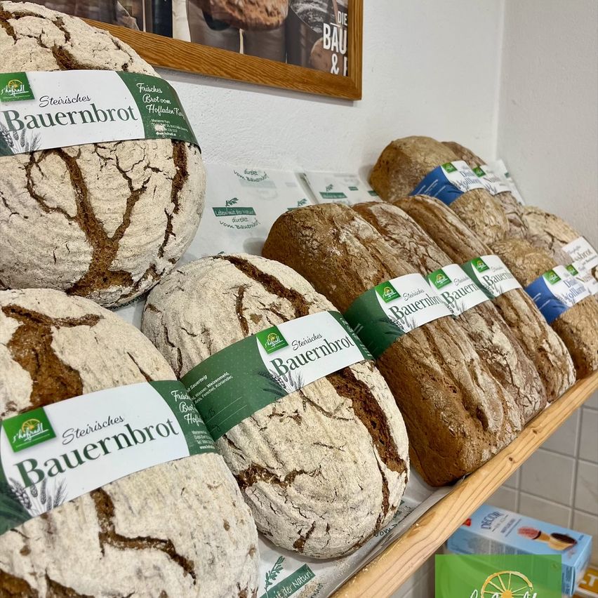 Ein Holzregal zeigt mehrere Laibe rustikales Brot, jeder in grünes und weißes Papier eingewickelt. Die Laibe sind mit 'Steirisches Bauernbrot' beschriftet und stehen auf einer weiß gefliesten Wand.