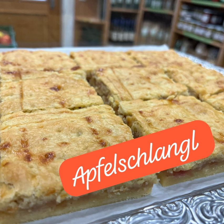 Ein Tablett mit Apfelscheiben, gebacken in einem goldbraunen Teig, auf einem glänzenden metallischen Tablett platziert. Das Wort 'Apfelschlangl' ist in Rot auf dem Bild geschrieben.