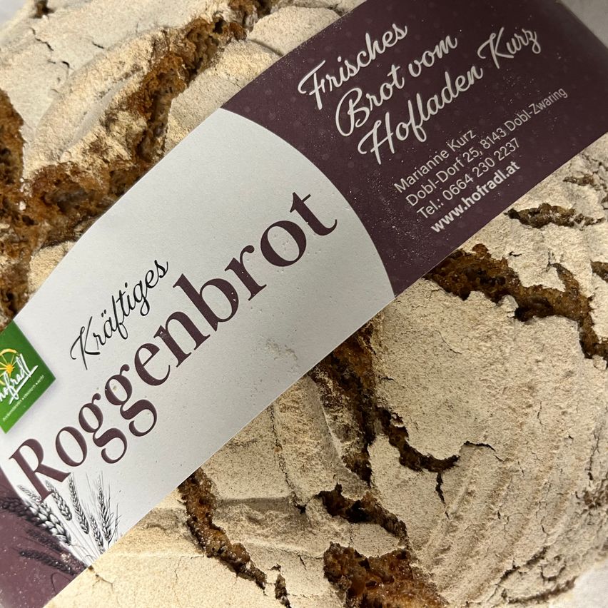 A close-up view of a bread loaf with a label reading 'Kraftiges Roggenbrot'. The label features a wheat drawing and a website address.