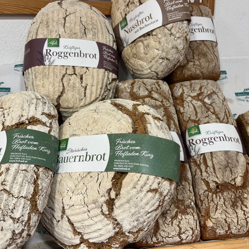 Ein Stapel von handwerklich hergestellten Broten, eingewickelt in Papier, einschließlich Roggenbrot, Nussbrot und Bauernbrot, auf einem Holzregal ausgestellt.