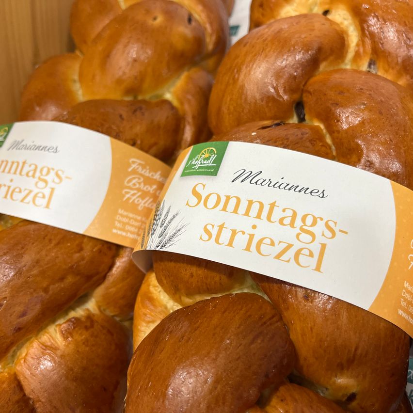 Eine Nahaufnahme mehrerer geflochtener Brotlaiben mit Etiketten, die 'Sonntags-striezel' in einer Bäckerei lesen. Das Brot hat eine goldbraune Kruste und ist leicht glänzend.