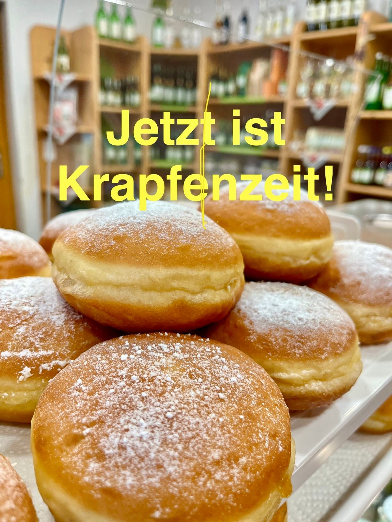 Ein Stapel frisch gebackener Donuts, die mit Puderzucker bestreut sind. Im Hintergrund befinden sich Regale mit verschiedenen Flaschen. Das Schild 'Jetzt ist Krapfenzeit!' zeigt an, dass es Zeit für Donuts ist.