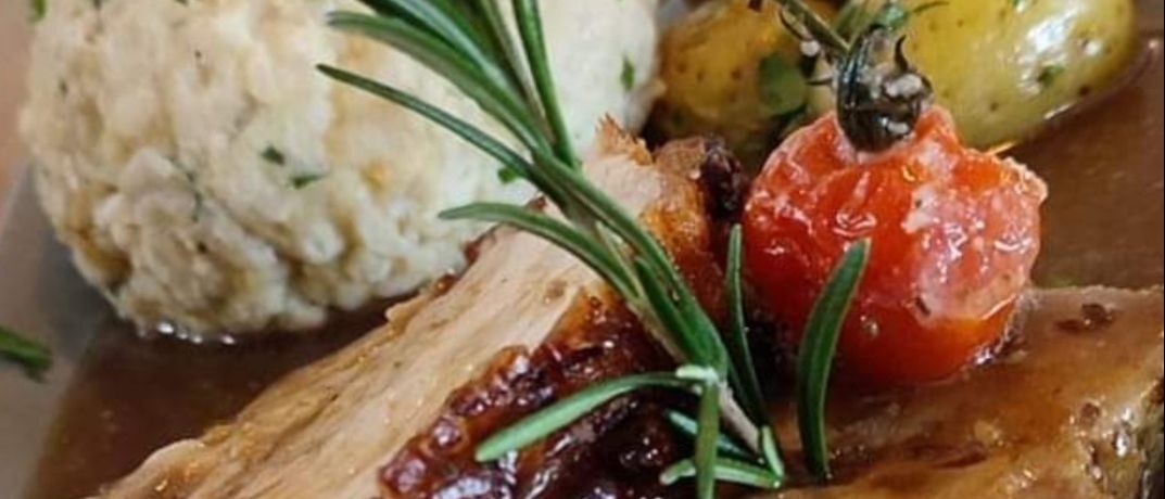 Ein Schweinefleischgericht mit glänzender Glasur, serviert mit Kartoffelpüree, einer Kirschtomate und einem Rosmarinzweig auf einem Teller.