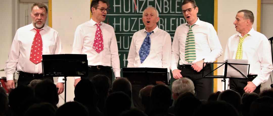 Drei Männer mit Krawatten singen auf der Bühne vor einem Publikum. Hinter ihnen zeigt eine Tafel einen Namen und das Wort 'HUZENBLEIBE'.