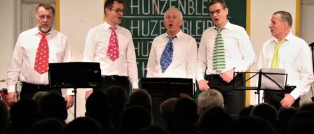 Drei Männer singen auf der Bühne mit Notenständern vor einer Tafel, auf der 'Huzenbleiber' steht, und einem Publikum.