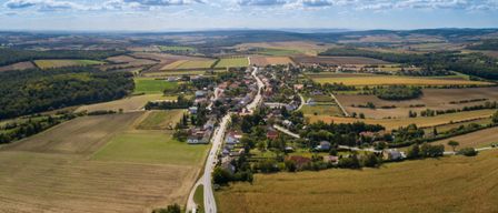 Luftaufnahme eines kleinen ländlichen Dorfes mit Feldern, Häusern und einer Straße unter einem teilweise bewölkten Himmel. Die Landschaft ist größtenteils flach mit grünen und braunen Feldern.