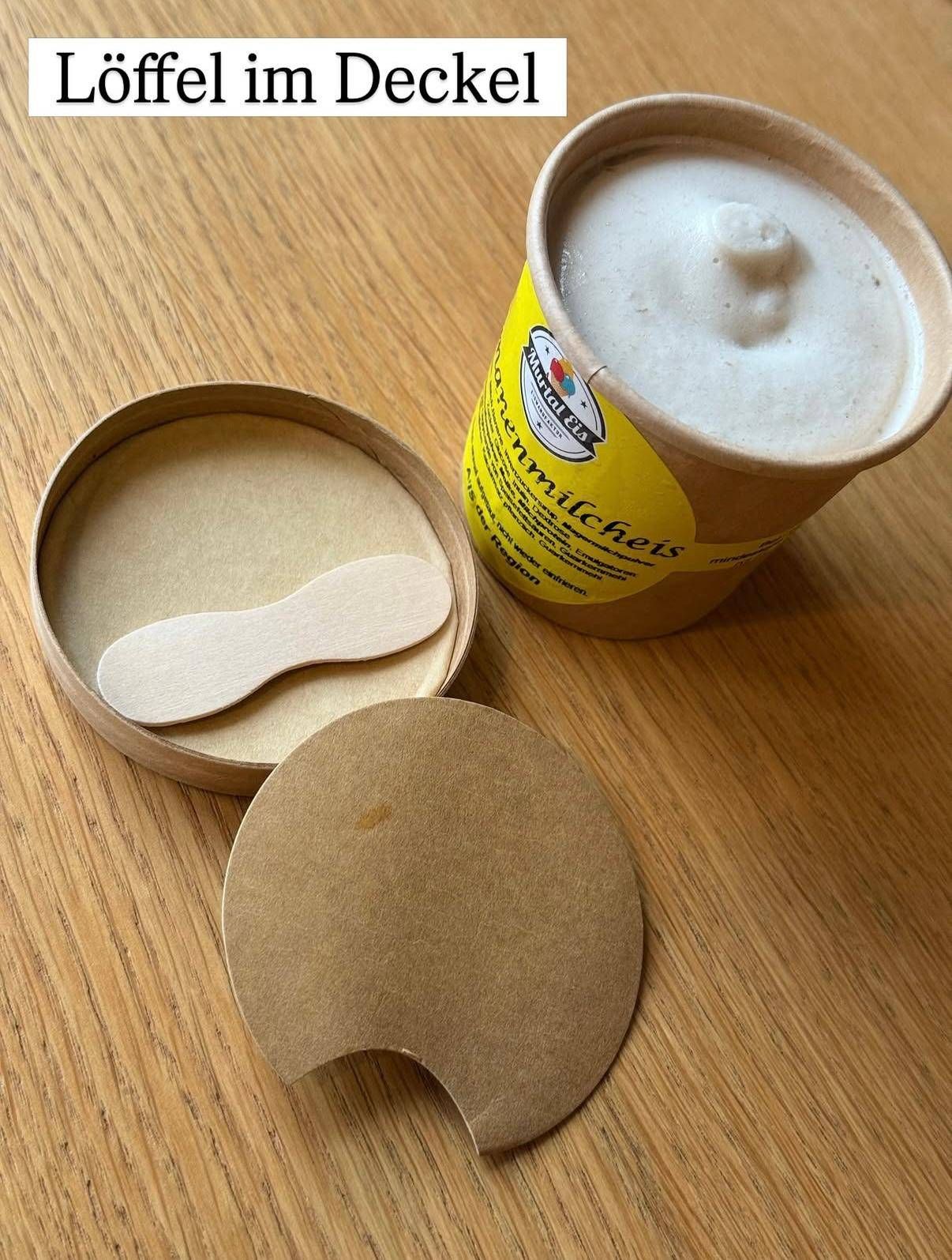 Ein hölzerner Löffel liegt in einer kreisförmigen Holzbox mit Deckel, neben einer Tasse Eiscreme mit gelbem Etikett.