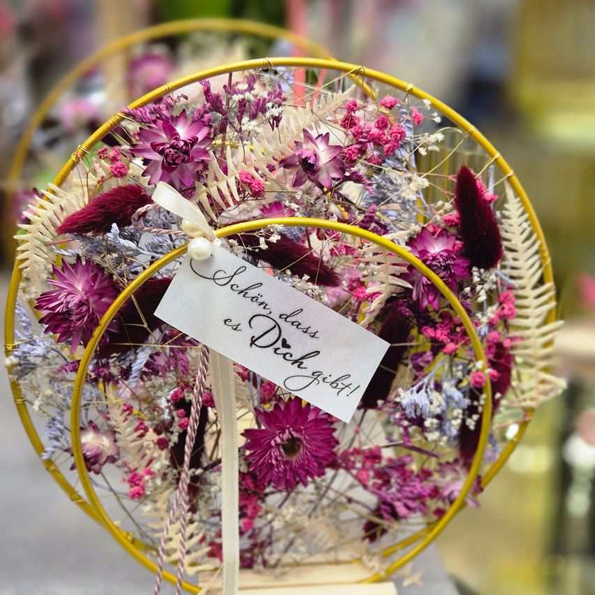 Eine kreisförmige Anordnung von getrockneten Blumen in Lila-, Rosa- und Weißtönen, mit einem gelben Rahmen und einem weißen Etikett, auf dem 'Schön dass es Dich gibt!' in kursiver Schrift steht.