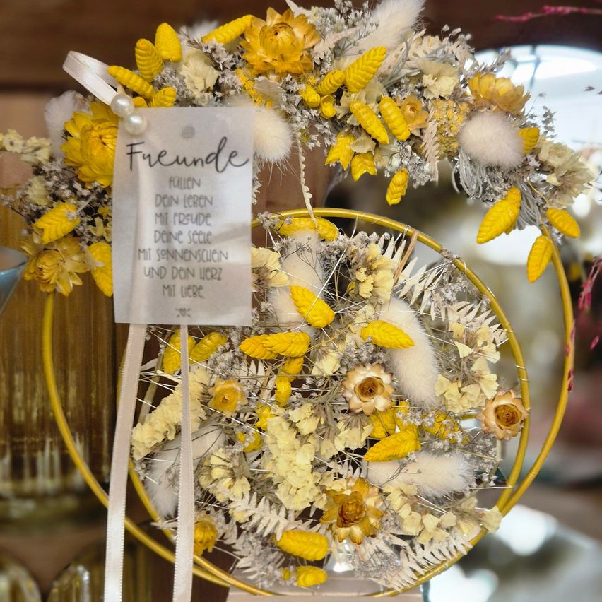 Ein gelber Kranz mit getrockneten Blumen und einer weißen Schleife wird in einem Geschäft ausgestellt. Ein kleiner Karton mit dem Text 'Freunde' ist am Kranz befestigt. Der Kranz steht auf einem gelben kreisförmigen Ständer.