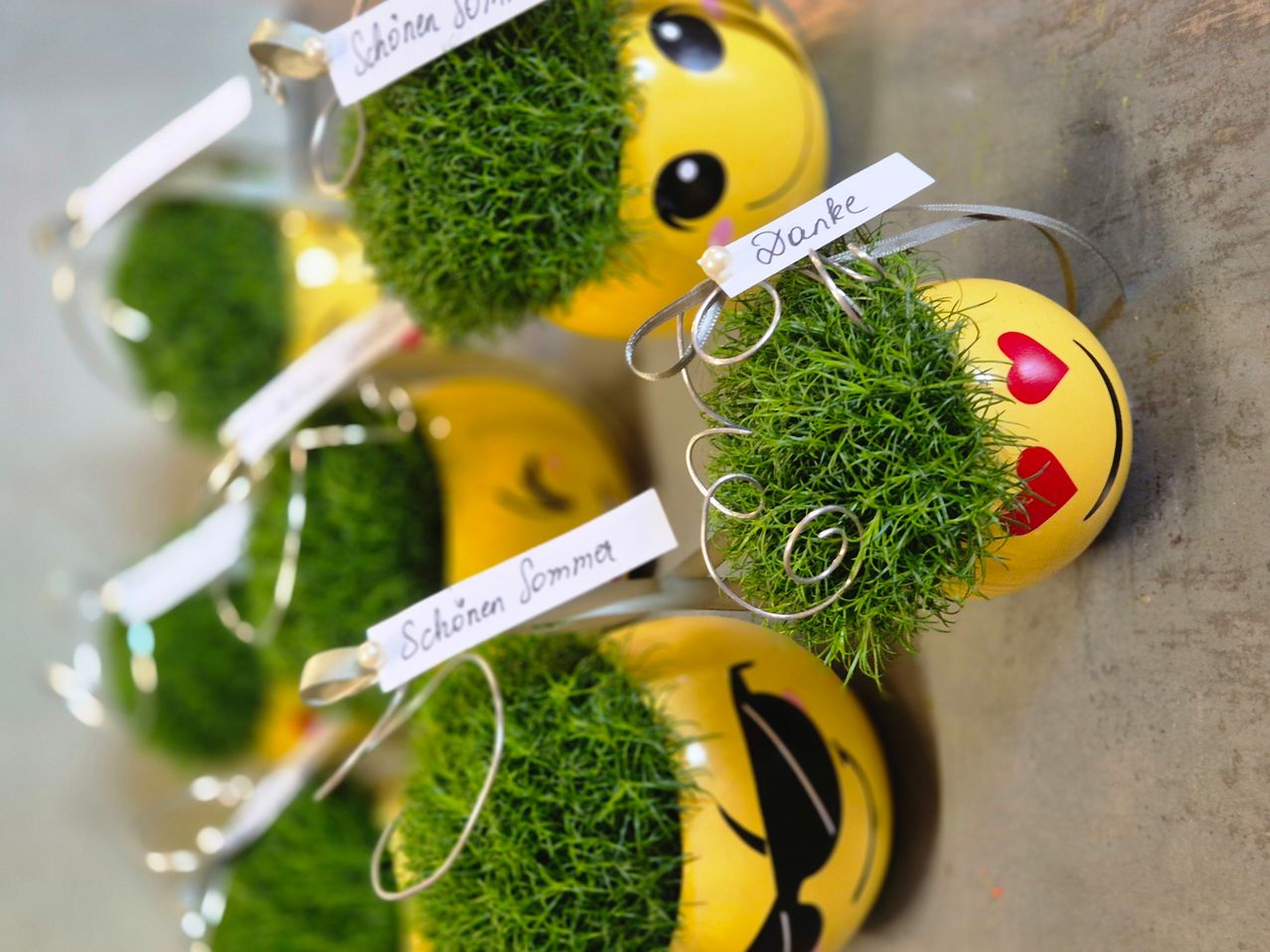 Several yellow spherical decorations with grass tops hang on a wall. Each has a face with expressions and a name tag. The tags read 'Schönen Sommer' and 'Danke'.