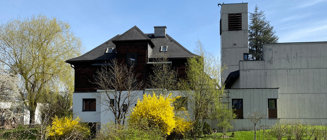 Ein Haus mit einem dunkelbraunen Dach und weißen Wänden steht vor einigen Bäumen und Büschen. Der Himmel ist klar.