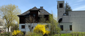 Ein Haus mit einem dunkelbraunen Dach und weißen Wänden steht vor einigen Bäumen und Büschen. Der Himmel ist klar.
